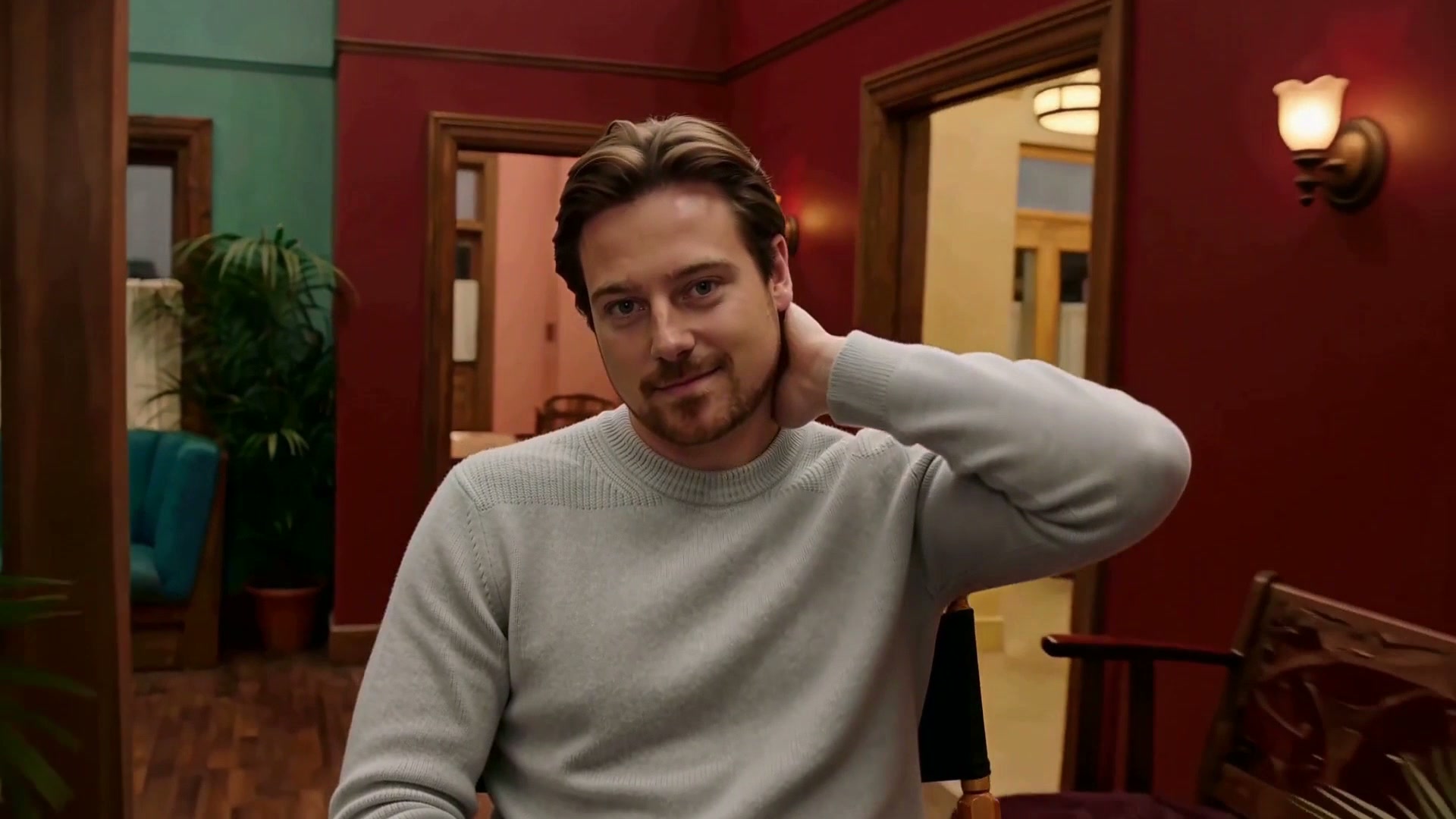 Replacement person in a pale sweater repeating the hand-to-neck gesture in the red room